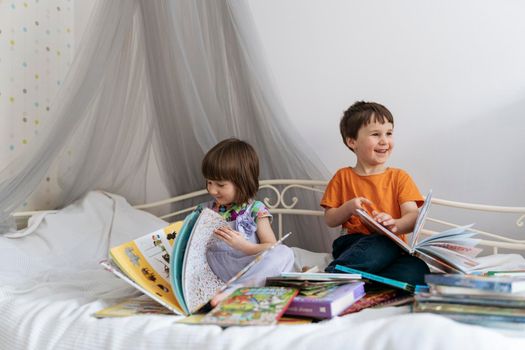 Two siblings reading books together on the sofa bed covered with white blanket in the kids' room, while both of them are smiling