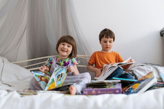 Two siblings reading books together on the sofa bed covered with white blanket in the kids' room, one of them smiling into the camera