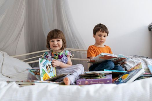 Two siblings reading books together on the sofa bed covered with white blanket in the kids' room, one of them smiling into the camera