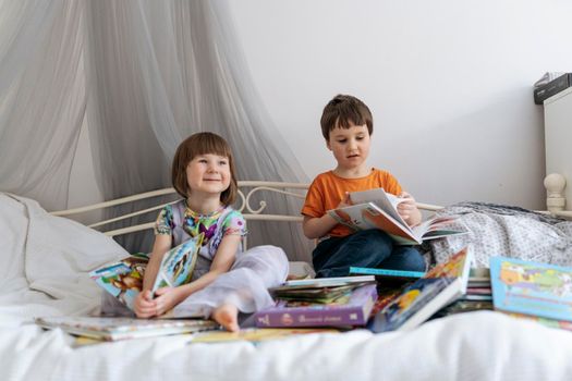 Two siblings reading books together on the sofa bed covered with white blanket in the kids' room, while both of them are smiling