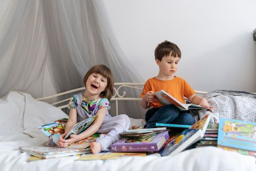 Two siblings reading books together on the sofa bed covered with white blanket in the kids' room, one of them laughing into the camera