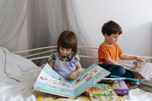 Two siblings reading books together on the sofa bed covered with white blanket in the kids' room