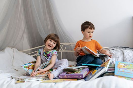 Two siblings reading books together on the sofa bed covered with white blanket in the kids' room, one of them laughing into the camera