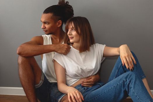 Multiracial family, stylish guy and girl in white t-shirts and blue jeans are sitting on the floor at home against a gray wall looking away.