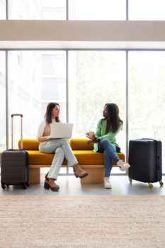 Young women waiting in hotel reception with trolley suitcases, talking, working with laptop. Vertical image. Lifestyle and travel concept.