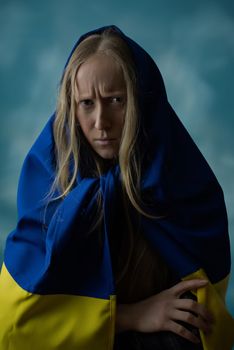 A real Ukrainian blonde woman is upset by evil and disappointed during the war with the state yellow and blue Ukrainian flag on her head. Russia attacked Ukraine on February 24, 2022.