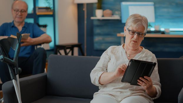Old woman using digital tablet with touch screen in living room. Senior person with crutches on couch browsing internet on device to have fun and enjoy retirement at home. Modern person