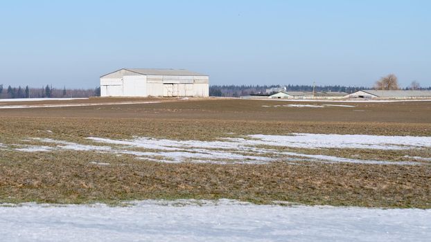 Melting field with dry grass and snow on the background of farm buildings.