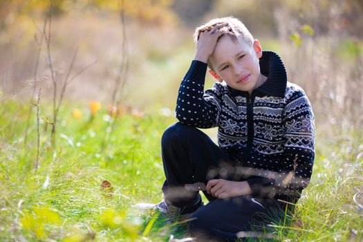 A handsome calm boy in a knitted sweater sits on the grass on the field.