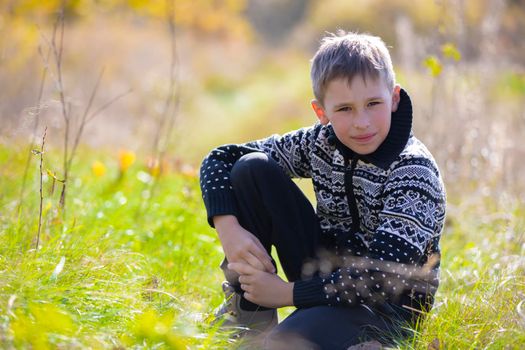 A handsome calm boy in a knitted sweater sits on the grass on the field.