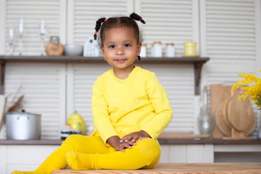 A little African American girl in a yellow dress with curly pigtails sits in the kitchen.
