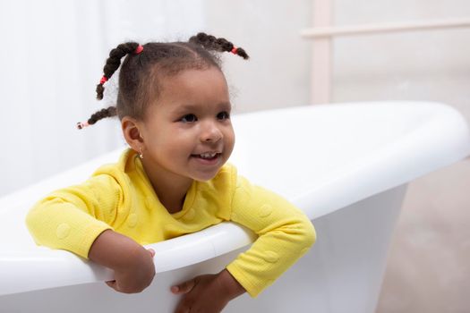 Little African American girl in a yellow dress with curly pigtails sits in a retro bath.