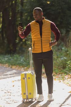 Cheerful african american young man in stylish clothes with suitcase walks in an autumn park on warm sunny day. Countryside travel concept