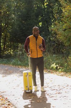 Cheerful african american young man in stylish clothes with suitcase walks in an autumn park on warm sunny day. Countryside travel concept