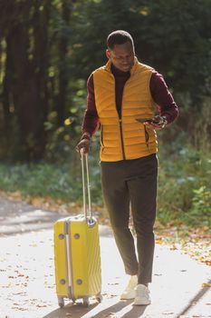 Cheerful african american young man in stylish clothes with suitcase walks in an autumn park on warm sunny day. Countryside travel concept