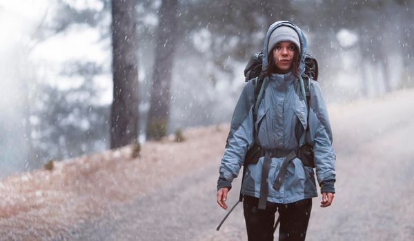 A young happy girl with hiking equipment and a backpack walks along the paths, in the mountains under the snowfall. The woman leads an active lifestyle and travels through the wilderness.