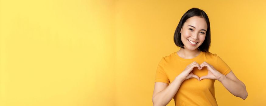 Beautiful asian girl showing heart, love gesture and smiling white teeth, express care and sympathy, standing over yellow background.