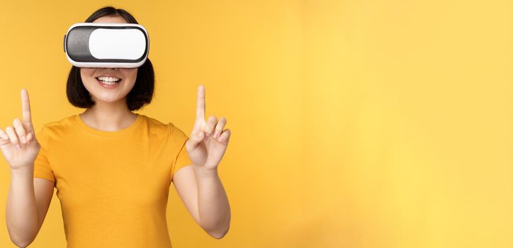 Young asian woman playing in VR, using virtual reality glasses, wearing yellow t-shirt against studio background. Copy space