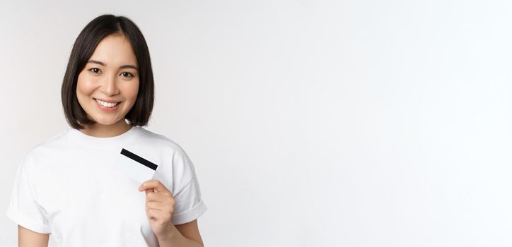 Portrait of korean smiling girl, bank client, showing credit card with happy face, standing against white background.