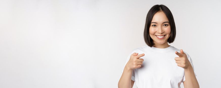 Image of smiling asian girl pointing fingers at camera, choosing, inviting you, congratulating, standing in tshirt over white background.