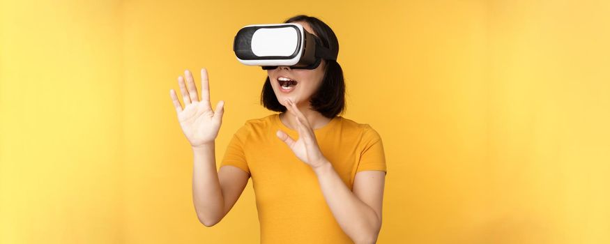 Girl in VR. Beautiful young asian woman, using virtual reality glasses and playing, chatting virtually, standing over yellow background.