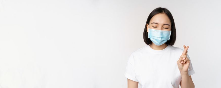 Covid-19, healthcare and medical concept. Image of asian girl in medical face mask, cross fingers, praying, making wish and smiling, standing over white background.