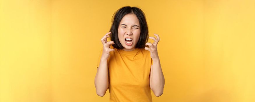 Image of angry asian woman, shouting and cursing, looking outraged, furious face expression, standing over yellow background.