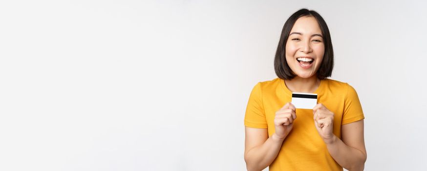 Portrait of beautiful korean girl holding credit card, recommending bank service, standing in yellow tshirt over white background.