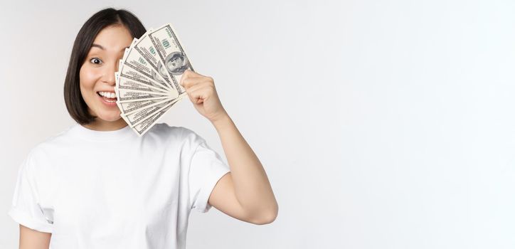 Portrait of smiling asian woman holding dollars money, concept of microcredit, finance and cash, standing over white background.