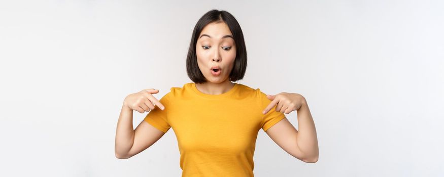 Happy asian girl pointing fingers down, looking at announcement, banner or advertisment, standing in yellow tshirt over white background.