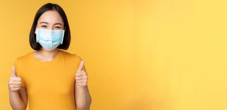 Smiling asian woman in medical face mask, showing thumbs up approval, like and recommend smth, standing over yellow background.