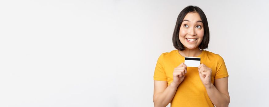 Portrait of beautiful korean girl holding credit card, recommending bank service, standing in yellow tshirt over white background.