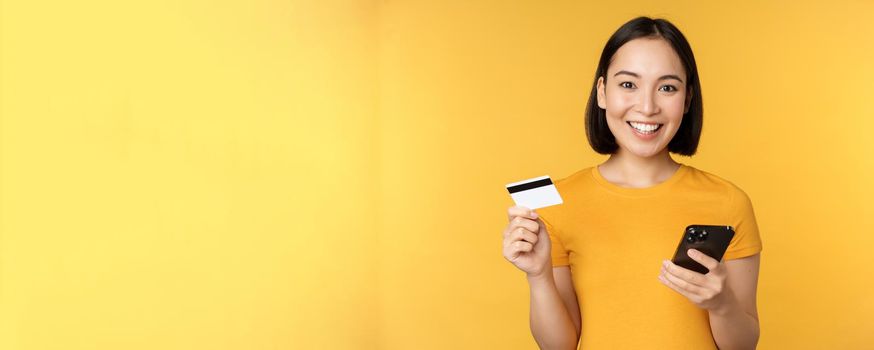 Online shopping. Cheerful asian girl holding credit card and smartphone, paying, order with mobile phone, standing over yellow background.