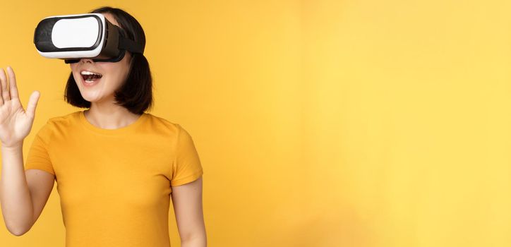 Girl in VR. Beautiful young asian woman, using virtual reality glasses and playing, chatting virtually, standing over yellow background.