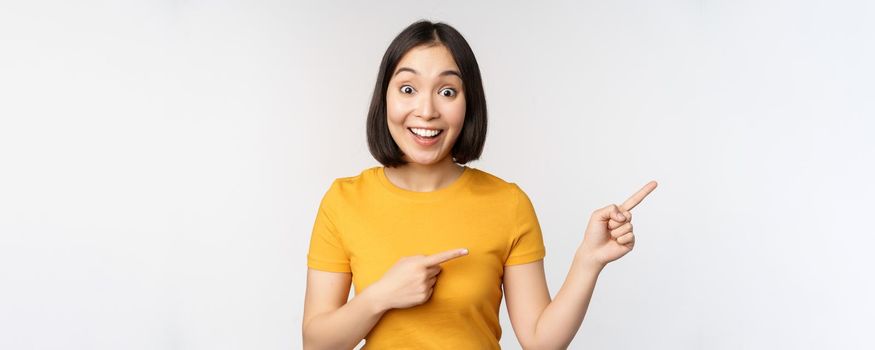 Amazed young asian woman, showing advertisement aside, pointing fingers right at promotion text, brand logo, standing happy against white background.
