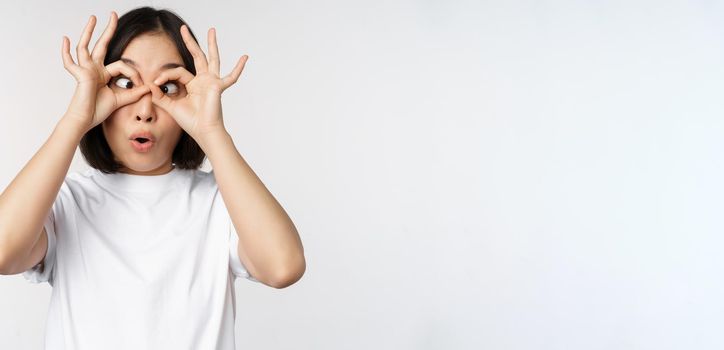 Funny young asian woman, korean girl making eyes glasses gesture, looking happy at camera, standing over white background.