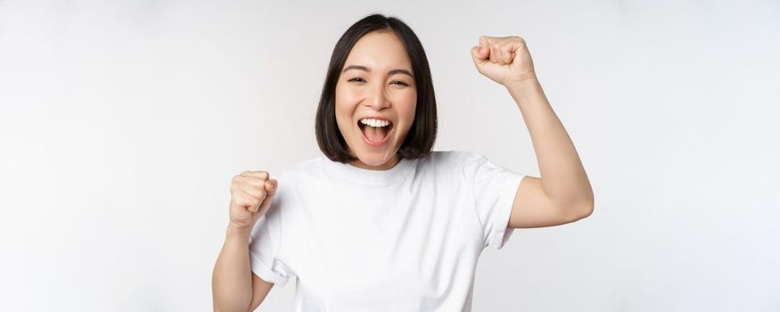 Portrait of enthusiastic asian woman winning, celebrating and triumphing, raising hands up, achieve goal or success, standing over white background.