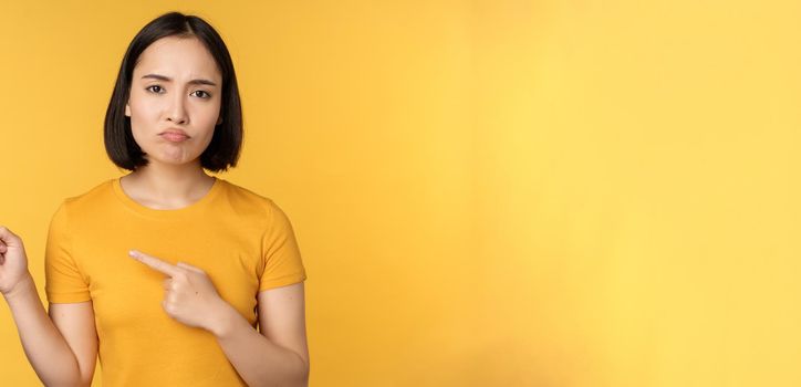 Upset asian woman pointing fingers left, showing smth disappointing, complaining at banner, standing over yellow background.