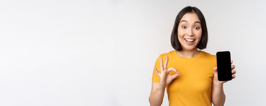 Excited asian girl showing mobile phone screen, okay sign, recommending smartphone app, standing in yellow tshirt over white background.