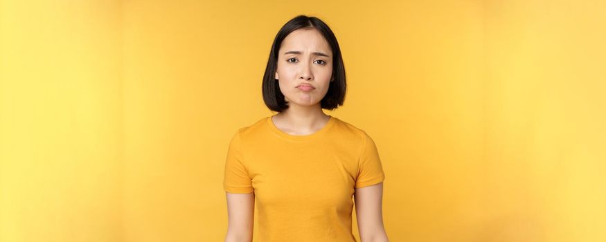 Disappointed asian girl sulking, looking upset, feel unair, standing in yellow t-shirt over white background. Copy space