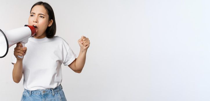 Asian girl shouting at megaphone, young activist protesting, using loud speakerphone, standing over white background. Copy space