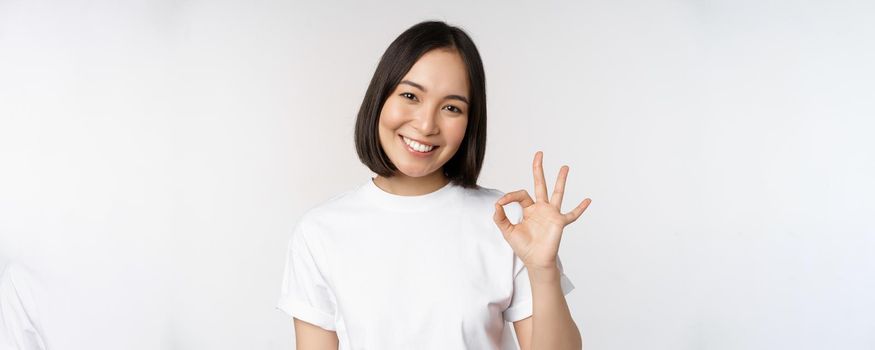 Everything okay. Smiling young asian woman assuring, showing ok sign with satisfied face, standing over white background. Copy space