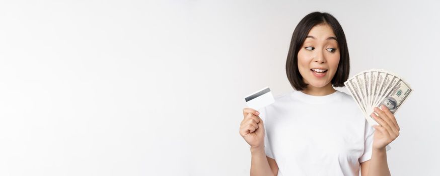 Portrait of asian woman holding money, dollars and credit card, looking impressed and amazed, standing in tshirt over white background.