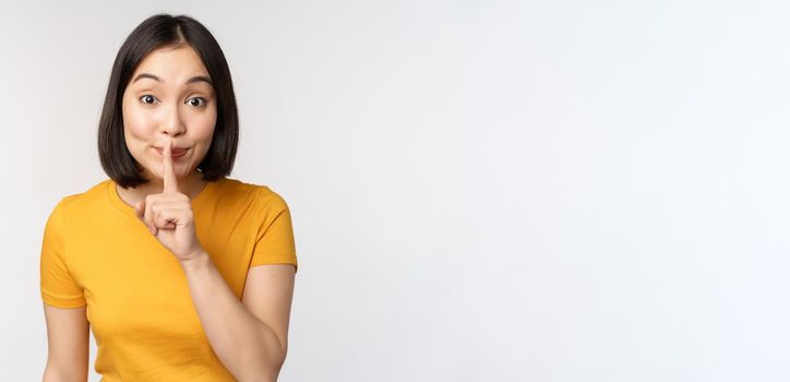 Keep quiet. Cute asian woman make shhh gest, showing shush, hush sign, press finger to lips, silence, standing over white background.