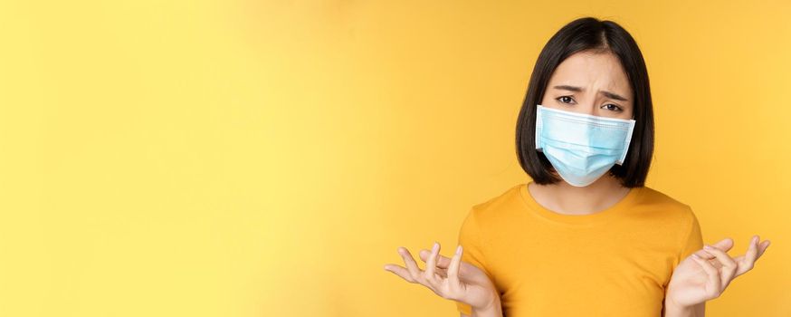 Close up portrait of confused asian woman in medical face mask, shrugging shoulders and looking puzzled, standing against yellow background.