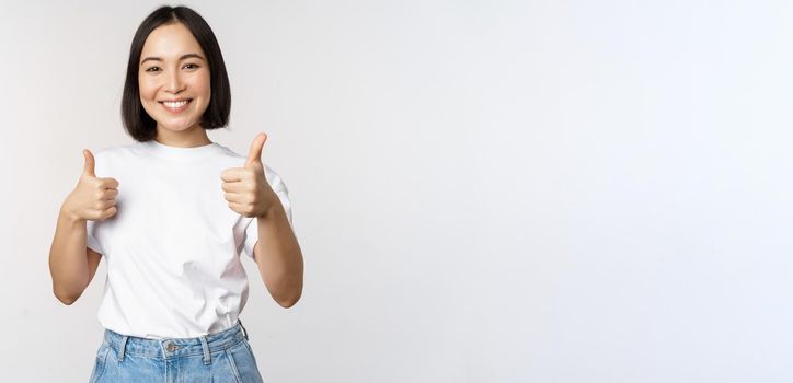 Happy beautiful korean girl, showing thumbs up in approval, smiling pleased, like smth, recommending, standing over white background.