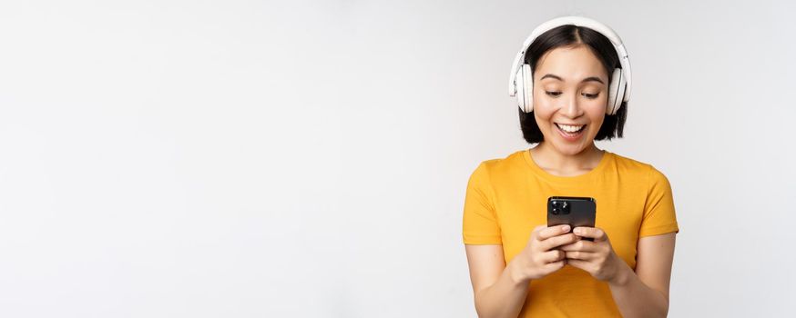 Cute japanese girl in headphones, looking at mobile phone and smiling, using music app on smartphone, standing against white background.