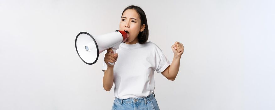 Asian girl shouting at megaphone, young activist protesting, using loud speakerphone, standing over white background. Copy space