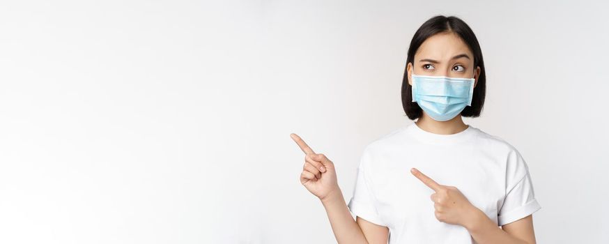 Image of asian woman in medical mask from covid, looking confused left, pointing at logo, showing advertisement, standing over white background.