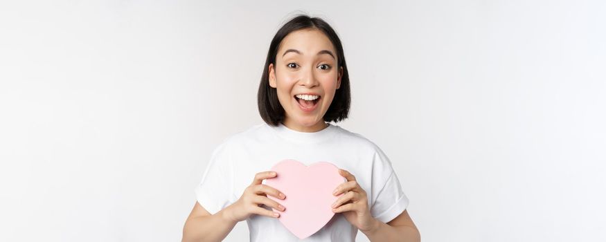 Valentines day. Happy asian girl receive romantic gift, holding heart shaped box and smiling excited, standing over white background.